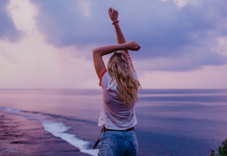 Back view anonymous female in denim shorts and white top standing on sandy beach with arms raised and enjoying picturesque view of evening seascape and purple sky