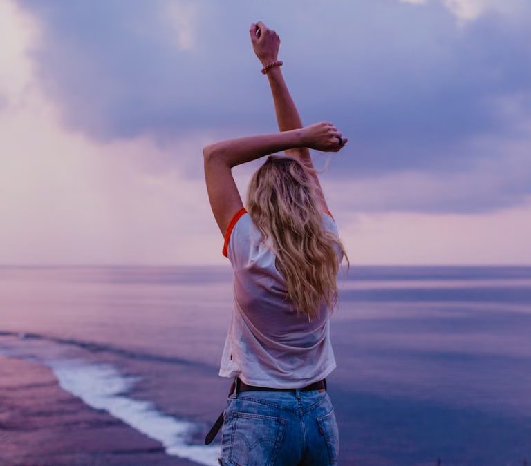 Back view anonymous female in denim shorts and white top standing on sandy beach with arms raised and enjoying picturesque view of evening seascape and purple sky
