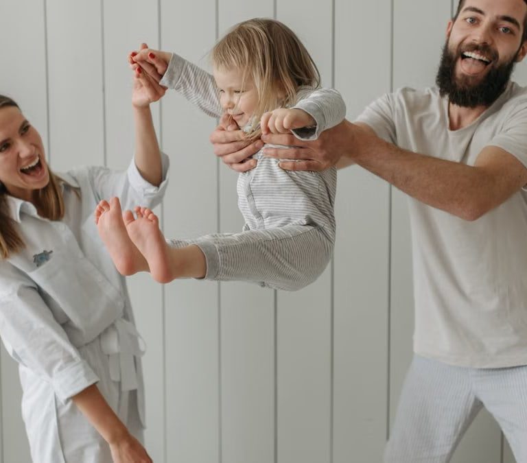Joyful parents playing with their daughter indoors, all wearing white, capturing a moment of happiness.